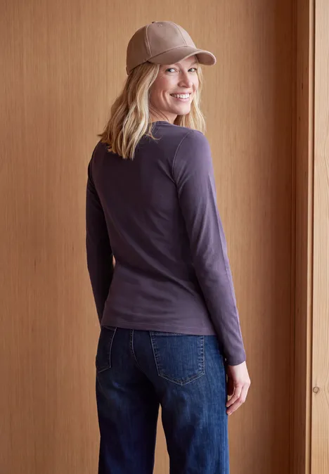 Langarm Shirt in Unifarbe mit V-Neck lilac shadow Eine junge Frau in Jeans und Cap steht vor Holzpaneelen und lächelt über die Schulter.