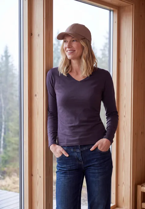 Langarm Shirt in Unifarbe mit V-Neck lilac shadow Lächelnde Frau mit Cap, dunkelblauem Shirt und Jeans steht in Holzhütte vor großem Fenster mit Waldblick.