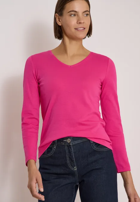 Langarm Shirt in Unifarbe mit V-Neck pink flash Frau in pinkem V-Ausschnitt-Pullover und dunklen Jeans, die lächelnd in die Ferne schaut.