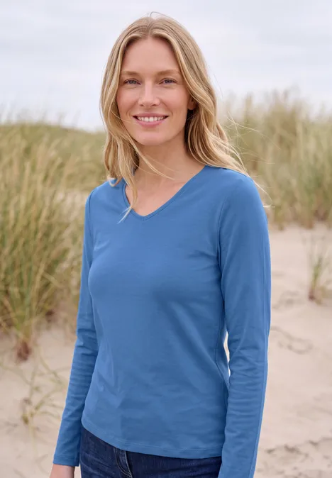 Langarm Shirt in Unifarbe mit V-Neck north blue Eine blonde Frau in blauem Langarmshirt steht am Strand vor Dünengras und lächelt.
