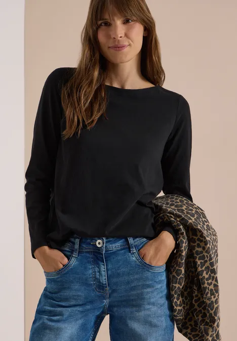 Kobieta w czarnym longsleeve i niebieskich jeansach nonszalancko trzyma na ramieniu wzorzystą kurtkę.