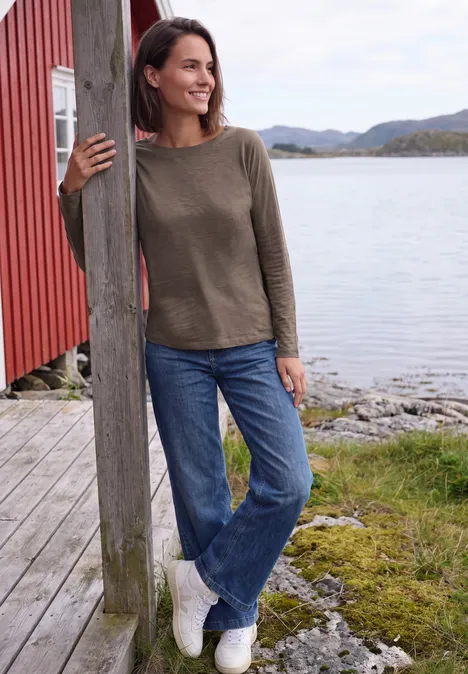 Shirt met lange mouwen en een boothals urban taupe Frau lehnt lächelnd an einem Holzpfosten am Seeufer, trägt Pullover und Jeans.