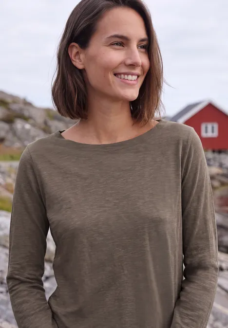 Shirt met lange mouwen en een boothals urban taupe Lächelnde Frau in olivgrünem Pullover, im Hintergrund Felsen und rote Häuschen an der Küste.