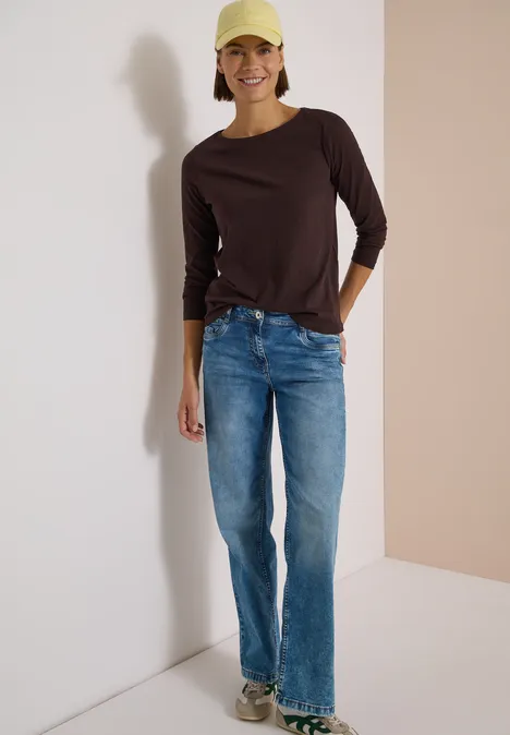 Langarm Shirt mit U-Boot-Ausschnitt truffle brown Frau mit gelber Schirmmütze, braunem Langarmshirt und Jeans lehnt locker an einer Wand und lächelt.