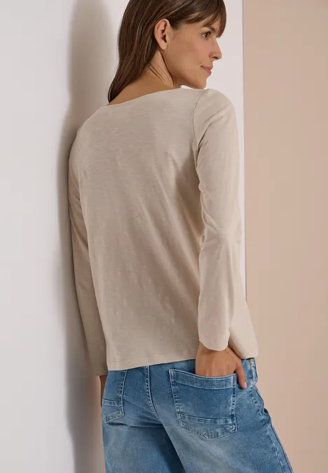 Eine Frau in hellbeigem Langarmshirt und blauen Jeans lehnt von hinten an einer hellen Wand.