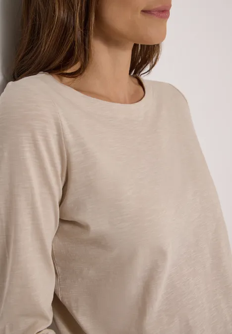 Frau mit beigem Langarmshirt lehnt sich an eine weiße Wand.