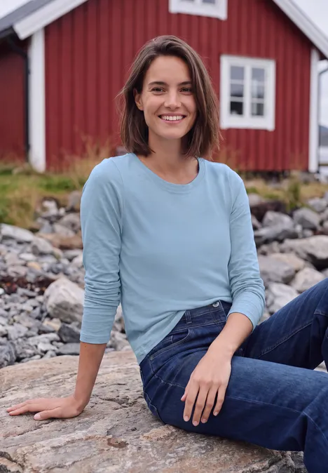 Langarm Shirt mit U-Boot-Ausschnitt cosmopolitan blue Eine lächelnde Frau in Jeans sitzt auf Kies vor einem rot gestrichenen Haus.