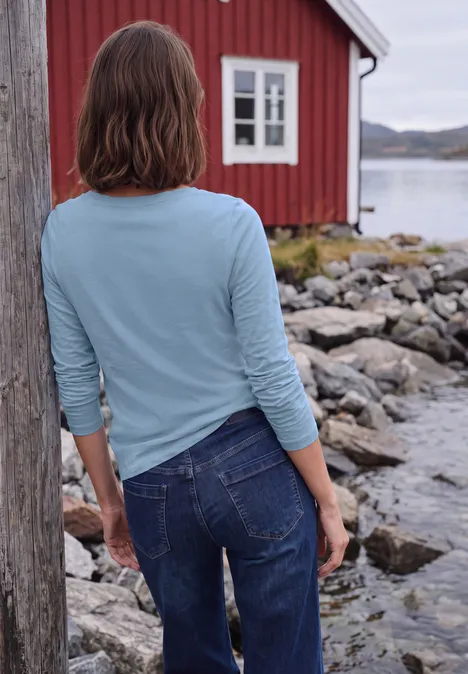 Langarm Shirt mit U-Boot-Ausschnitt cosmopolitan blue Person mit blauer Bluse steht mit dem Rücken zur Kamera vor einem roten Haus am felsigen Wasserufer.