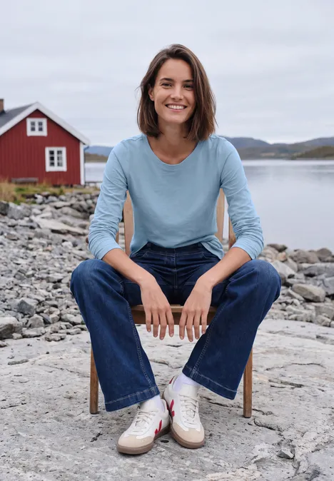 Langarm Shirt mit U-Boot-Ausschnitt cosmopolitan blue Eine lächelnde Frau in Jeans und blauem Shirt sitzt auf einem Stuhl am felsigen Ufer mit rotem Haus im Hintergrund.