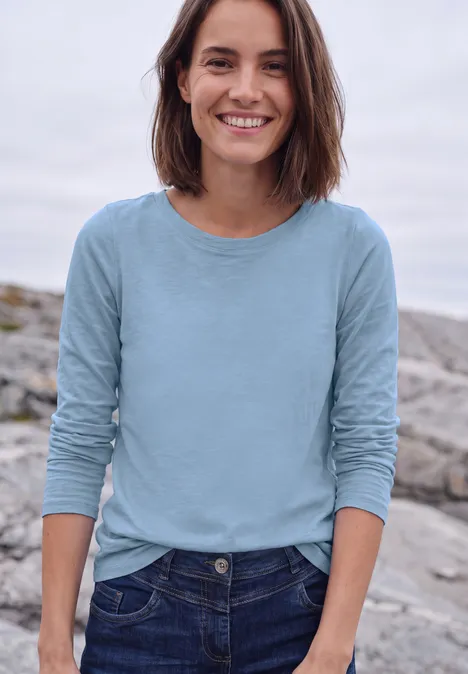 Langarm Shirt mit U-Boot-Ausschnitt cosmopolitan blue Eine lächelnde Frau in Jeans und hellblauem Langarmshirt steht auf Felsen am Wasser.