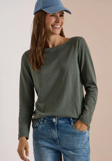 Frau trägt grünes Longsleeve, Jeans und blauen Cap, lächelt.