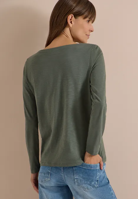 Frau mit langen braunen Haaren in olivgrünem Langarmshirt, Rückenansicht, Hände in Jeans-Taschen.