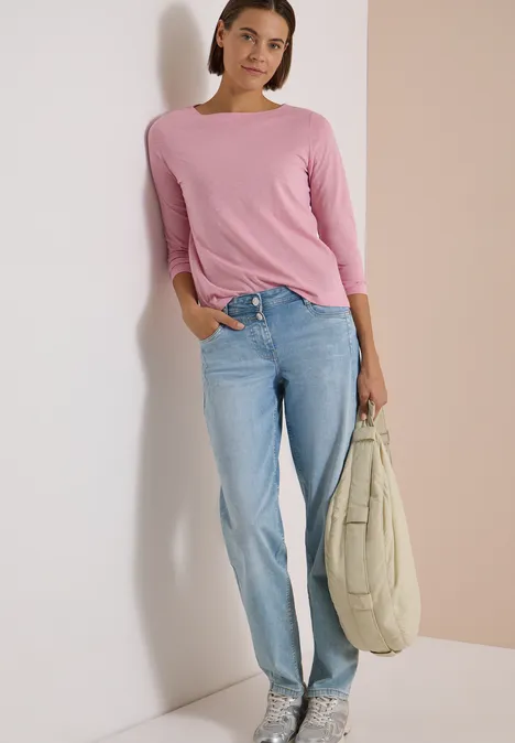 Langarm Shirt mit U-Boot-Ausschnitt mellow rose Frau in pinkem Shirt und blauen Jeans lehnt lächelnd an der Wand und hält eine große Tasche.