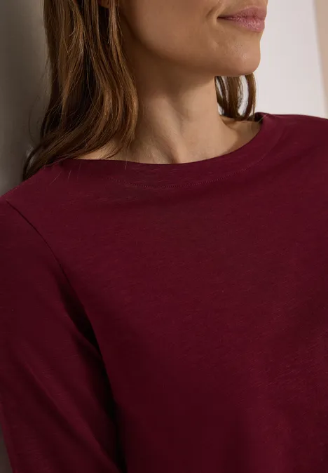 Teilaufnahme einer Frau mit burgunderfarbenem Langarmshirt und leicht seitlich gedrehtem Oberkörper.