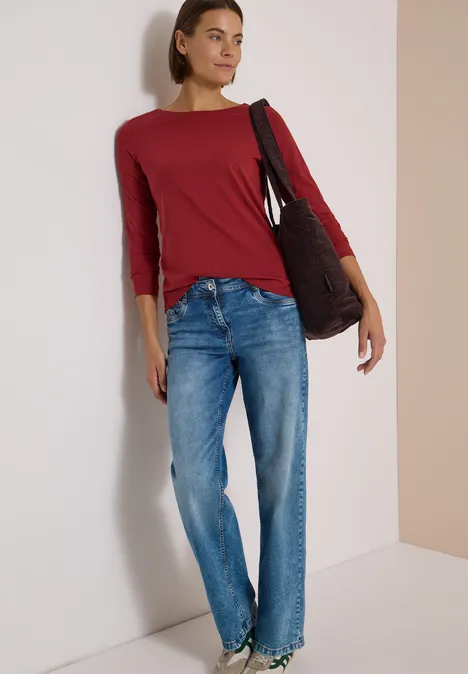 Langarm Shirt mit U-Boot-Ausschnitt russet red Eine Frau in rotem Langarmshirt, blauen Jeans und Tasche steht lässig an einer Wand.