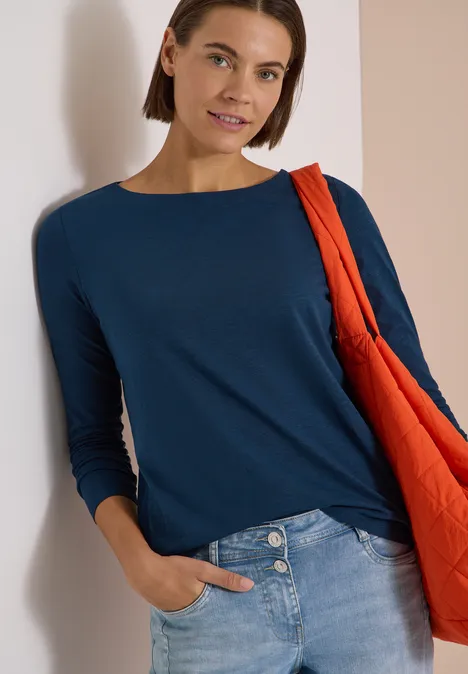 Langarm Shirt mit U-Boot-Ausschnitt moonlight blue Frau in dunkelblauem Langarmshirt, Jeans und orangener Stepp-Tasche lehnt an einer Wand.