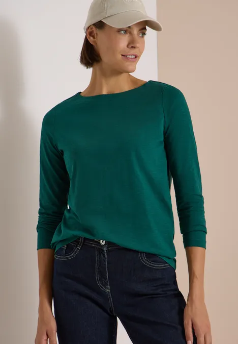 Langarm Shirt mit U-Boot-Ausschnitt dark fir green Eine Frau trägt ein grünes Langarmshirt, blaue Jeans und eine weiße Baseballkappe.