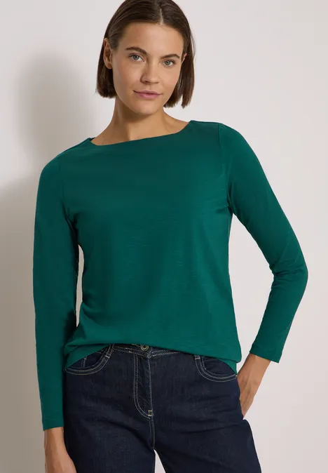 Langarm Shirt mit U-Boot-Ausschnitt dark fir green Frau mit grünem Langarmshirt und dunkler Jeans vor hellem Hintergrund.