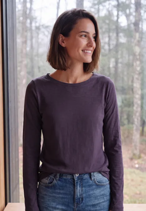 Langarm Shirt mit U-Boot-Ausschnitt lilac shadow Eine lächelnde Frau mit braunem Bob, lila Longsleeve und Jeans steht am Fenster vor Wald