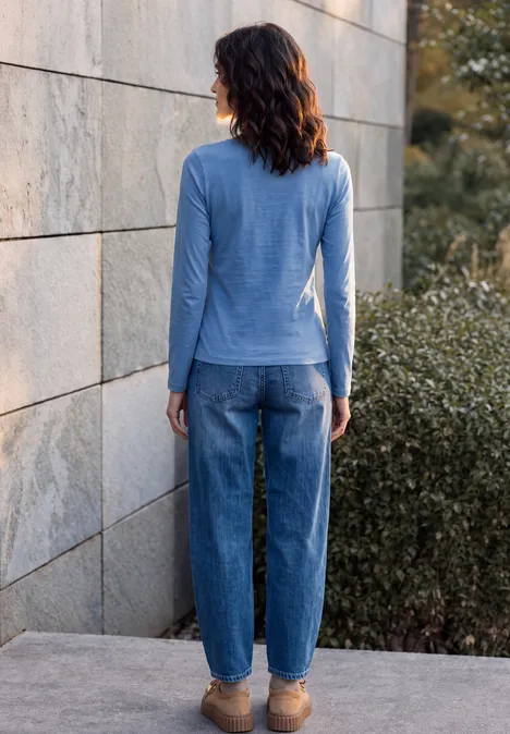 Frau von hinten in blauem Langarmshirt und Jeans, steht vor einer Steinmauer im Freien.