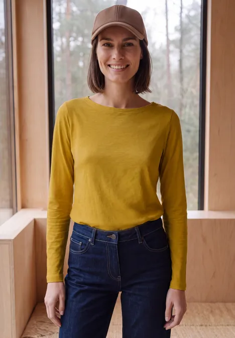 Eine Frau mit braunem Hut, gelbem Langarmshirt und Jeans steht lächelnd vor einem Fenster in einem Holzraum.
