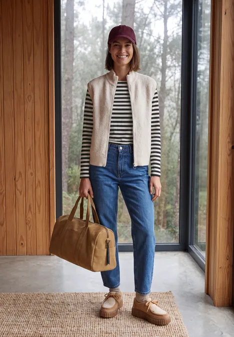 Langarm Shirt mit Boat Neck und Streifen Black Frau steht lässig in Jeans, gestreiftem Shirt, Strickweste, Mütze und trägt eine Reisetasche vor Glasfenster.