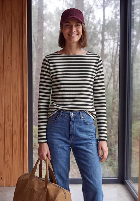 Langarm Shirt mit Boat Neck und Streifen Black Frau in gestreiftem Shirt, Jeans, lila Cap hält braune Tasche vor großem Fenster.