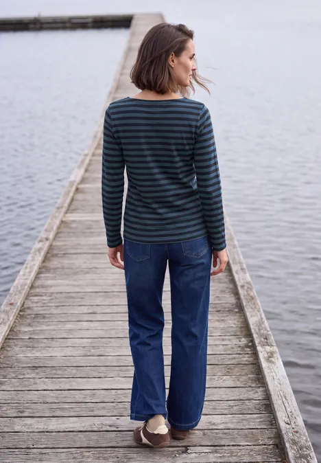 Frau in gestreiftem Pullover und Jeans steht auf einem Holzsteg am Wasser, Blick nach links.
