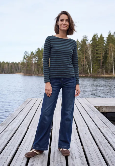 Frau steht auf Holzsteg am See, trägt gestreiftes Shirt und Jeans.