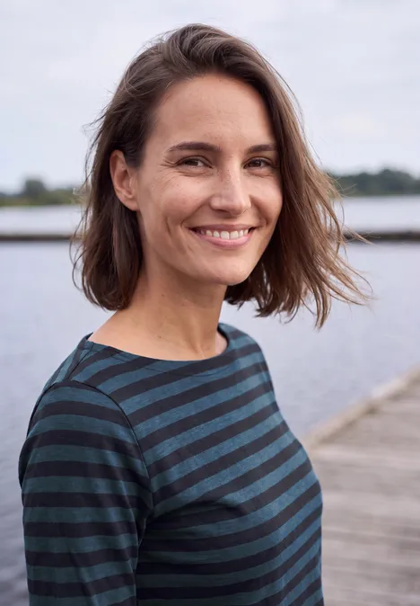Eine lächelnde Frau mit kurzen braunen Haaren und gestreiftem Oberteil steht auf einem Steg am Wasser.