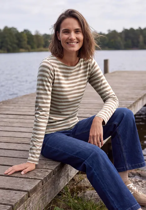 Een lachende vrouw zit op een houten steiger aan een meer, in een gestreept shirt en spijkerbroek.