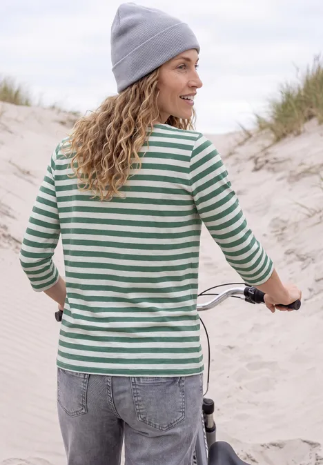 Frau in gestreiftem Shirt und Mütze schiebt Rad über sandige Dünenküste.