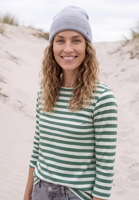 Eine lächelnde Frau mit gestreiftem Shirt und Mütze steht am Strand in den Dünen.