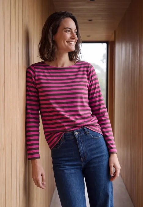 Vrouw glimlacht in een houten gang, draagt een roze-grijs gestreept shirt met lange mouwen en jeans.