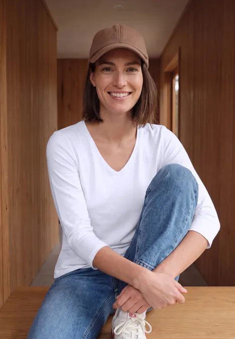 Eine lächelnde Frau in Jeans, weißem Oberteil und Basecap sitzt vor Holzwänden.
