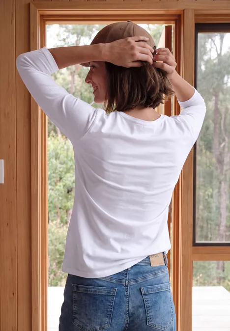 Frau von hinten in weißem Shirt und Jeans richtet ihre kurzen Haare in einem hellen Raum mit großen Fenstern.