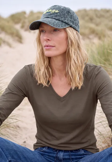Frau mit Basecap und grünem Shirt sitzt entspannt in einer Dünenlandschaft.