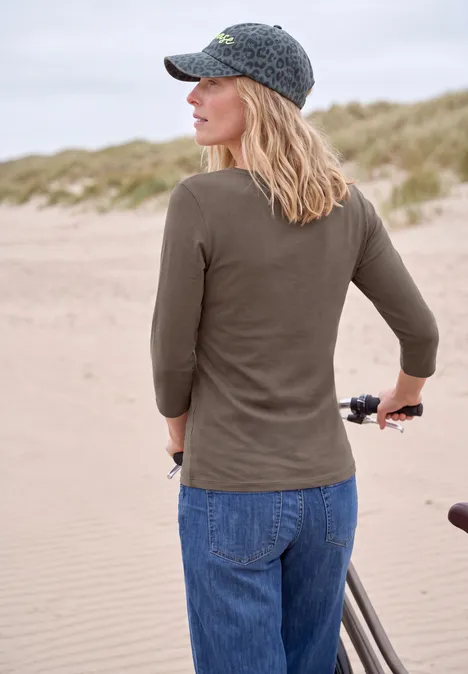 Frau in Kappe und Pullover schiebt ihr Fahrrad entlang sandiger Dünen am Strand.