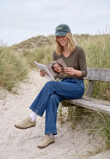 Frau sitzt auf einer Bank zwischen Dünen am Strand und liest eine Zeitschrift.