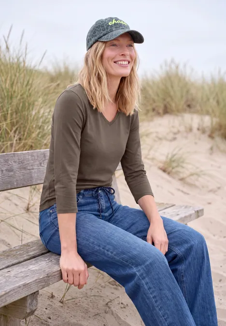 Frau sitzt lächelnd in Jeans und Mütze auf einer Strandbank, Dünengras im Hintergrund.