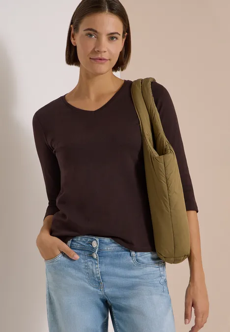 3/4-Arm Shirt in Unifarbe mit V-Neck truffle brown Frau in dunkelbraunem V-Shirt, hellblauen Jeans und olivgrüner gesteppter Tasche vor beigem Hintergrund.