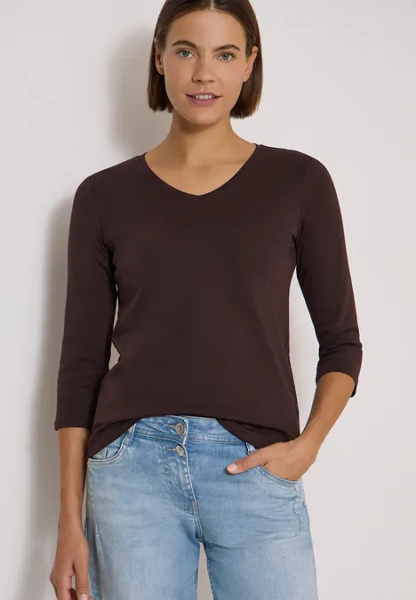 3/4-Arm Shirt in Unifarbe mit V-Neck truffle brown Frau in braunem Langarmshirt und blauen Jeans, Hand in Tasche, lächelnd vor hellem Hintergrund.