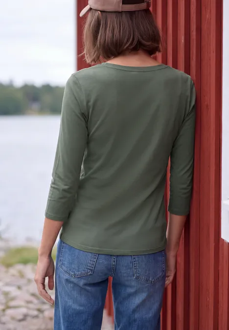 Iemand van achteren met groen 3/4-mouwshirt en spijkerbroek leunend tegen een rood houten muur bij een meer.