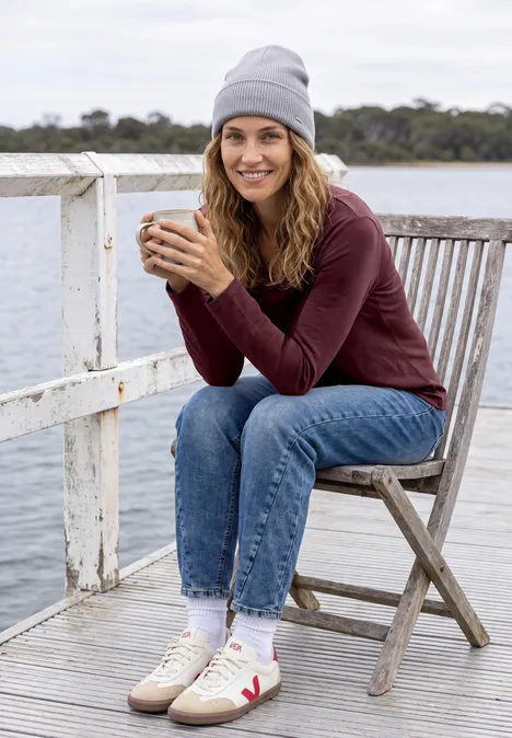 Eine lächelnde Frau sitzt auf einem Steg, hält eine Tasse, trägt Mütze, Pullover und Jeans.
