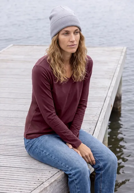 Frau in grauer Mütze, rotem Langarmshirt und Jeans sitzt nachdenklich auf einem Holzsteg am Wasser.