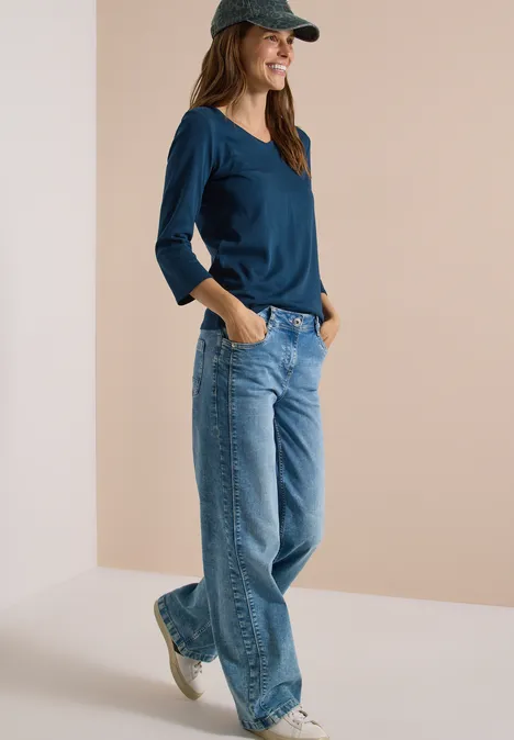 3/4-Arm Shirt in Unifarbe mit V-Neck moonlight blue Eine Frau in Jeans, blauem Langarmshirt und Cap, läuft lässig mit Händen in den Taschen.