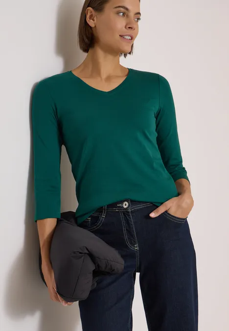 3/4-Arm Shirt in Unifarbe mit V-Neck dark fir green Frau in grünem V-Shirt, dunkelblauer Jeans und Jacke in der Hand vor neutralem Hintergrund.