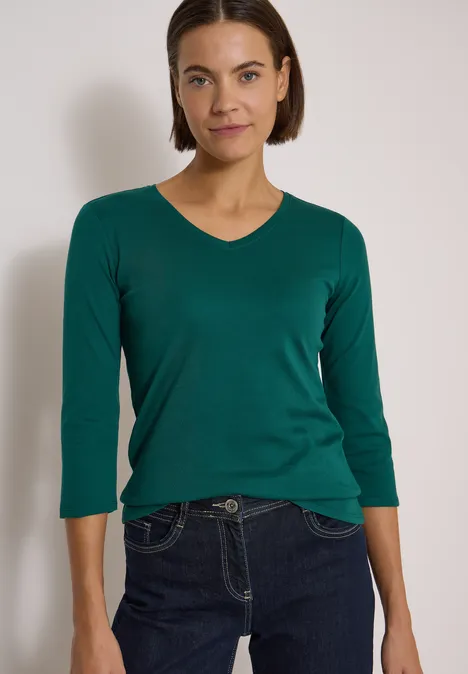 3/4-Arm Shirt in Unifarbe mit V-Neck dark fir green Frau mit grünem V-Ausschnittshirt (3/4-Ärmel) und dunkler Jeans vor neutralem Hintergrund.