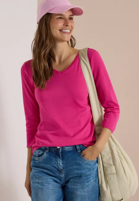 3/4-Arm Shirt in Unifarbe mit V-Neck pink flash Frau in pinkem Shirt, Jeans, Cap und Schultertasche lächelt vor neutralem Hintergrund.