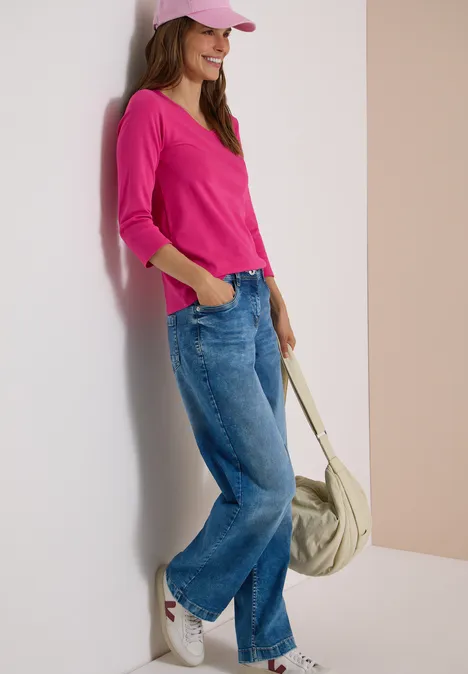 3/4-Arm Shirt in Unifarbe mit V-Neck pink flash Frau lehnt an Wand, trägt rosa Pullover und Cap, weite Jeans, hält lässig eine Tasche.